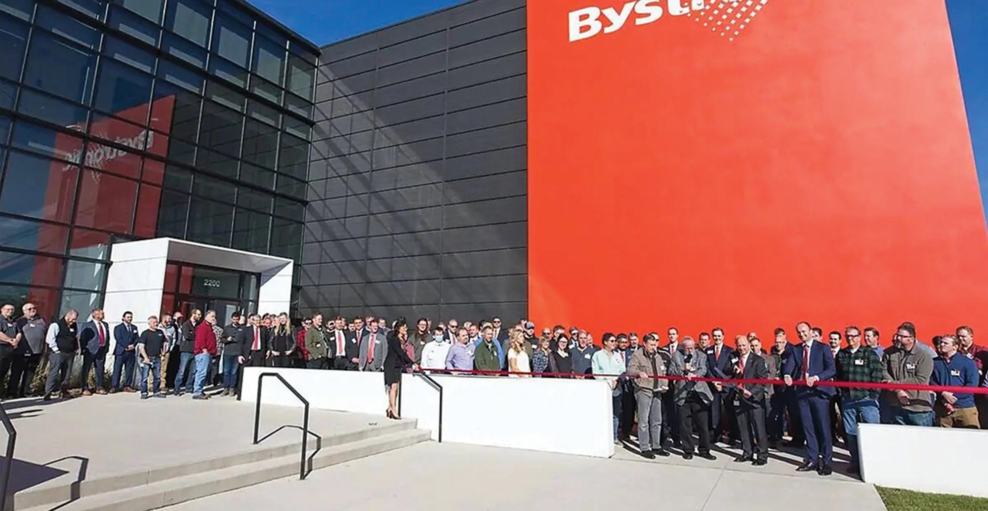 a large crowd stands behind red tape with a Bystronic Inc. building in the background